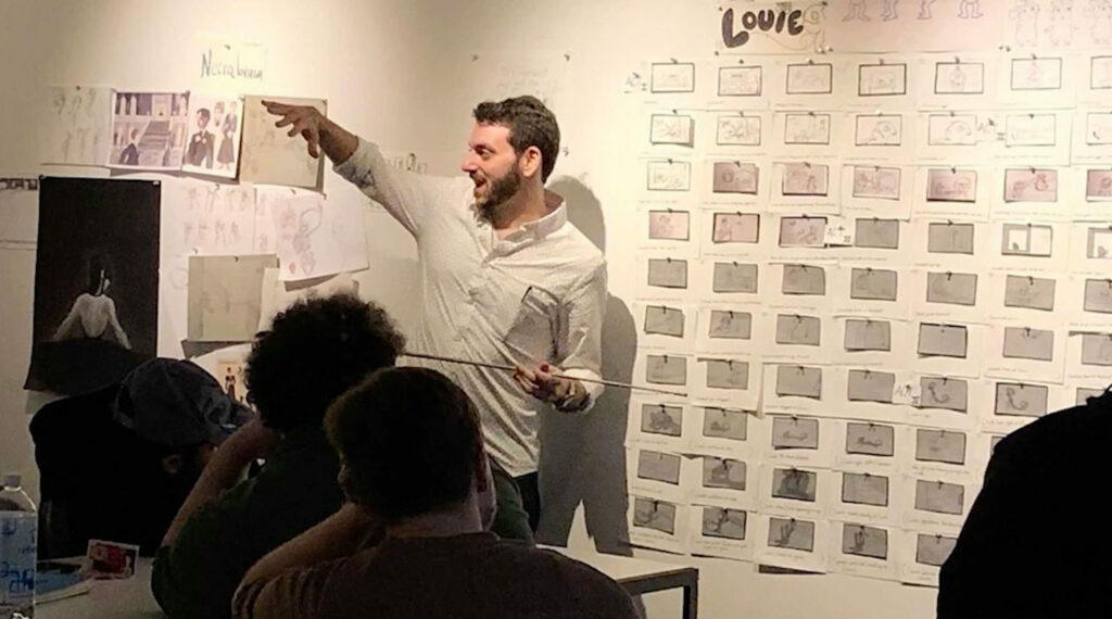 Instructor gestures with a pointer at a storyboard wall labeled "Louie." Students sit nearby in dimly lit art classroom with sketches and posters surrounding them.